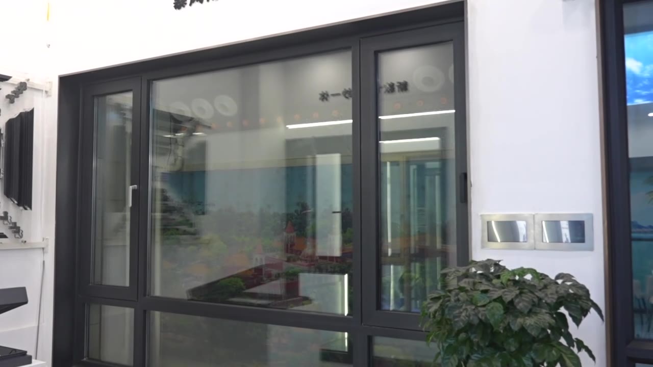 Full-size black-framed aluminium sample window with a fixed centre light and two opening sashes mounted in a white sample wall, with the glazing reflecting the room behind the camera