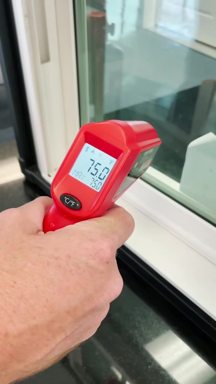 Handheld infrared thermometer taking a surface temperature reading on a white uPVC window frame during factory performance testing